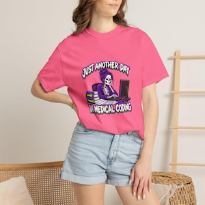 Op de afbeelding: Een felroze t-shirt met korte mouwen en een afbeelding van een skelet met paars haar dat aan een computerbureau met boeken zit. De tekst JUST ANOTHER DAY IN MEDICAL CODING staat in witte letters met een groene omlijning op de voorkant.