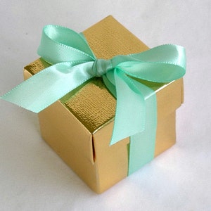 May include: A small, square gift box with a gold metallic finish. The box is tied with a light green satin ribbon and bow.