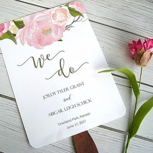 May include: A white wedding fan with a floral design at the top. The text "we do" is written in cursive script. The names "Jordy Tyler Grant" and "Abigail Leigh Schick" are printed below the text. The date "June 17, 2017" and the location "Overland Park, Kansas" are printed below the names.