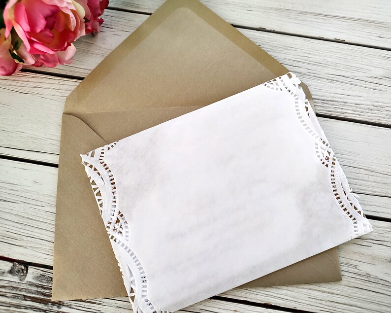 May include: A white card with a lace border is placed inside a brown envelope. The envelope and card are on a white wooden surface.