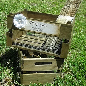 May include: A rustic wooden crate with a wire mesh insert, used to hold wedding programs. The crate is decorated with a white flower and a sign that reads "Programs John & Cindy June 12, 2019".