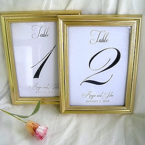 May include: Two gold-framed table number signs with black calligraphy. The signs read "Table 1" and "Table 2". The text "Angie and John August 2, 2018" is printed below the table numbers.