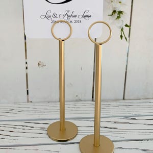 Op de afbeelding: Twee hoge goudkleurige metalen tafelnummerhouders met ronde voetstukken en ringvormige bovenkanten. Een witte kaart zit in de ringen, met de zwarte tekst 3, Liza & Andrew Lemon, en 23 MARCH, 2018, en een klein wit bloemaccent ernaast.