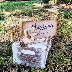 May include: A gold wire basket with a wooden handle sits on a whitewashed wooden crate. A sign on the basket reads "Programs" with the names "arrett & Nicole" and the date "10.14.23". A decorative flower is attached to the basket.