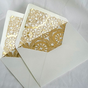 May include: Two white envelopes with ornate gold laser-cut patterns visible inside. The gold designs feature floral and circular shapes. The envelopes are open, showcasing the detailed gold embellishments.