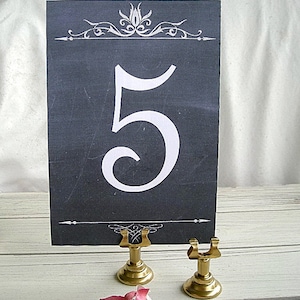 Può includere: Un segnatavolo rettangolare grigio scuro con un grande numero 5 bianco al centro, decorato con ghirigori bianchi nella parte superiore e inferiore, tenuto in posizione verticale da un piccolo supporto in metallo dorato con base circolare.