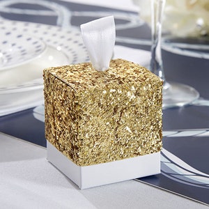 Op de afbeelding: Een klein vierkant doosje met een gouden glitterafwerking en een wit lint. Het doosje staat op een blauw-wit gepatroneerd oppervlak.