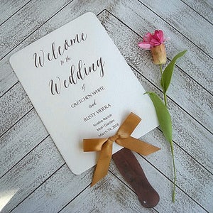 May include: White wedding fan with a gold ribbon and a wooden handle. The fan has the text "Welcome to the Wedding of Gretchen White and Rusty Vierra, Kualoa Ranch, Molii Garden, March 24, 2018".