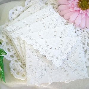 Puede incluir: Una pila de sobres blancos con bordes de encaje. Los sobres tienen un diseño delicado y calado y están dispuestos sobre un tapete blanco de ganchillo. Una flor rosa es visible en el fondo.