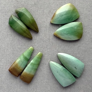 May include: Six polished gemstone pieces in shades of green, brown, and white are arranged on a gray surface. The shapes vary, including pointed and rounded forms, showcasing natural color variations and textures.