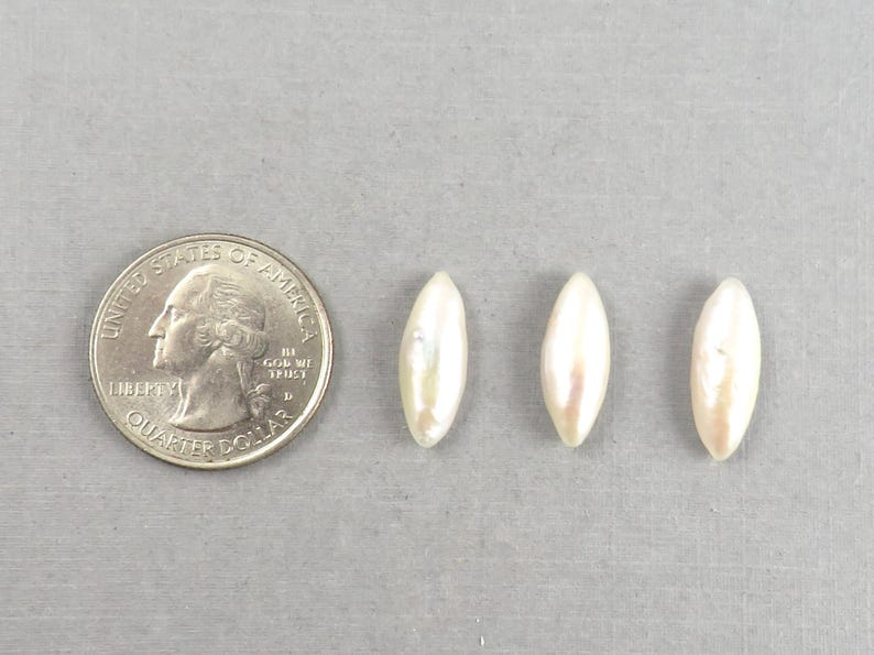 May include: Three lustrous, elongated, off-white pearls are displayed next to a U.S. quarter coin. The pearls have a subtle iridescent sheen. The quarter provides a size reference for the pearls, which are approximately 0.75 inches in length.