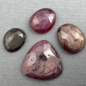 May include: Four faceted gemstones in shades of pink, brown, and gray. The stones are oval and triangular, with a textured surface. The gems are arranged on a gray surface.