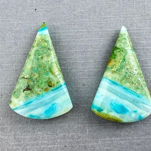 May include: Two triangular-shaped gemstones with a mix of green, blue, and gold hues. The stones have a layered appearance, with a band of turquoise blue separating the green and gold sections. The gemstones are set against a gray background.