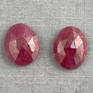 May include: Two faceted, oval-shaped, ruby gemstones. The gems are a deep red color and have multiple facets that catch the light. The stones are set against a gray background.