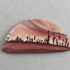 May include: A polished, semi-circular stone with a natural landscape pattern. The stone features bands of reddish-brown and cream colors, with a dark brown, tree-like pattern along the bottom edge. The stone is set against a gray background.