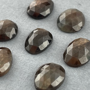 Pode incluir: Uma coleção de pedras preciosas ovais e facetadas em tons de castanho. As pedras têm uma superfície polida e refletora, e estão dispostas sobre um fundo de cor neutra. As facetas criam um jogo de luz e sombra.