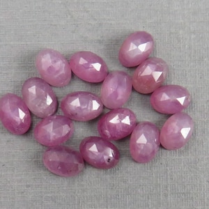 May include: A collection of oval-shaped, faceted gemstones in varying shades of pink. The gemstones are scattered on a light gray surface, with each stone displaying multiple facets that catch the light, creating a sparkling effect. The gems are likely rubies.