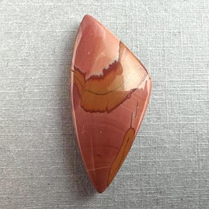 May include: A polished, teardrop-shaped gemstone with a blend of colors. The stone features a base of reddish-brown with layers of tan, orange, and white. The stone has a smooth, glossy surface and is set against a light gray background.
