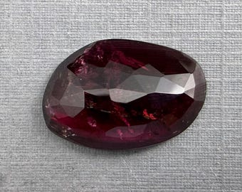 May include: A faceted, oval-shaped gemstone with a deep burgundy color. The gem has multiple facets that reflect light, creating a sparkling effect. The stone is set against a gray textured background.