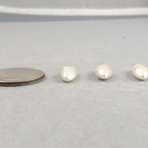 May include: A quarter coin next to three off-white, oval-shaped pearls. The pearls are smooth and have a slight luster. The coin is silver in color and has a ridged edge.