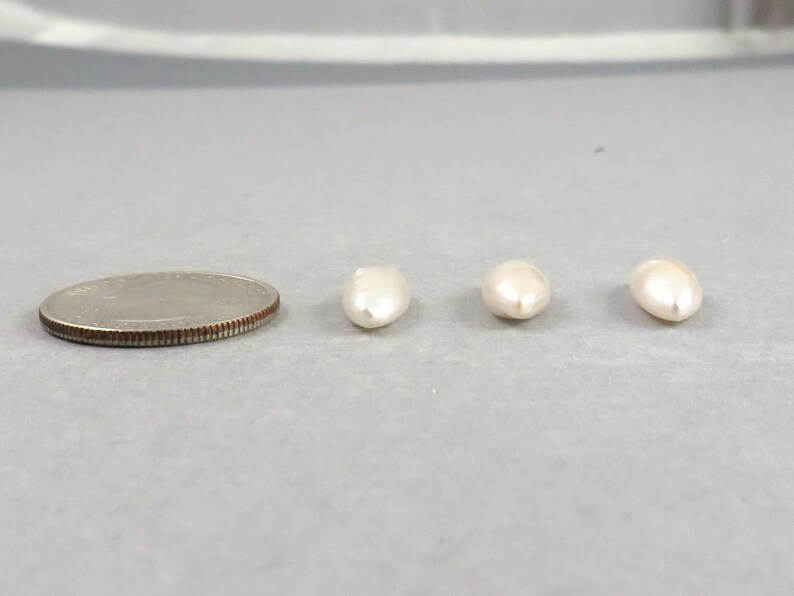 May include: A quarter coin next to three off-white, oval-shaped pearls. The pearls are smooth and have a slight luster. The coin is silver in color and has a ridged edge.