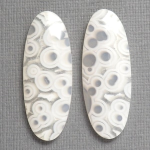 May include: Two oval-shaped gemstones with a white and gray pattern. The pattern consists of concentric circles and irregular shapes, creating a unique, organic design. The gemstones are set against a gray background.
