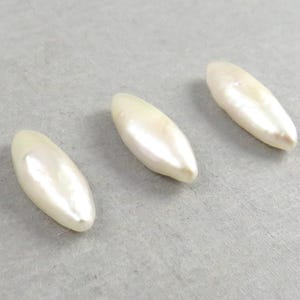 May include: Three elongated, off-white, pearlescent beads are arranged on a light gray surface. Each bead has a smooth, glossy finish and a slightly irregular shape, suggesting they are natural. The beads are evenly spaced.