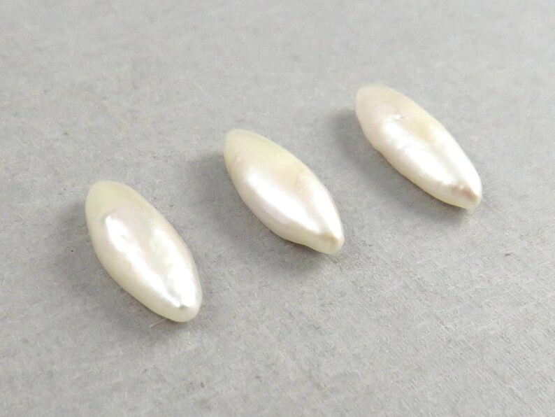 May include: Three elongated, off-white, pearlescent beads are arranged on a light gray surface. Each bead has a smooth, glossy finish and a slightly irregular shape, suggesting they are natural. The beads are evenly spaced.