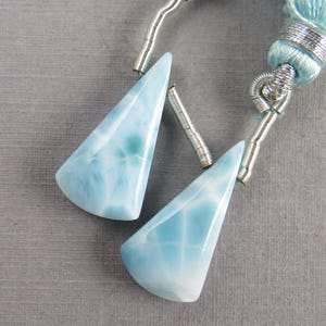 Könnte beinhalten: Zwei hellblaue, dreieckige Edelsteinanhänger mit marmoriertem Aussehen. Die Anhänger sind an silberfarbenen Metallfassungen und einer hellblauen Quaste befestigt. Die Edelsteine haben eine polierte, glänzende Oberfläche.