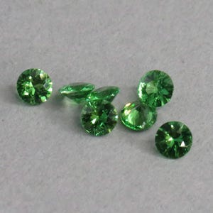 May include: A collection of round, faceted, bright green gemstones. The gems are scattered on a light gray surface, showcasing their brilliant cut and vibrant color. These gemstones could be used for jewelry making.