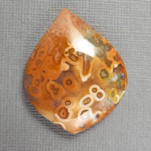 Può includere: Un cabochon di pietra preziosa lucidato a forma di goccia con un motivo complesso di inclusioni orbicolari in tonalità arancione bruciato, crema, marrone chiaro e marrone. La superficie è liscia e riflettente, con segni circolari naturali.