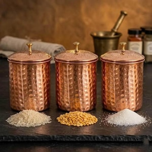 Op de afbeelding: Drie cilindrische koperen voorraadpotten met een gehamerde textuur en bijpassende deksels. Elke pot heeft een kleine knop als handvat op de bovenkant. De potten staan op een rij met kleine hoopjes witte rijst, gele linzen en wit zout.