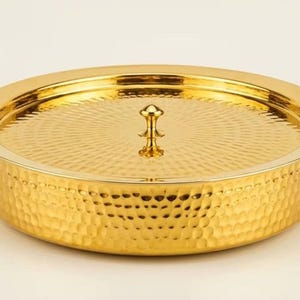 Op de afbeelding: Een ronde, goudkleurige metalen serveerschaal met bijpassend deksel. De buitenkant van de basis heeft een gehamerde textuur, terwijl het deksel een radiaal zonnestraalpatroon heeft. Een kleine centrale knop zit op het deksel en twee ringgrepen.