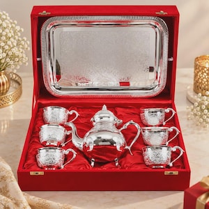 May include: A silver tea set featuring a teapot and four matching cups with intricate floral engravings, presented in a red velvet-lined box. A rectangular silver tray with a decorative border is mounted inside the lid of the storage case.