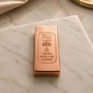 Pode incluir: Uma barra de cobre retangular com acabamento metálico polido. A parte frontal apresenta um design gravado com o símbolo químico Cu, um ícone de árvore e o texto 500 GRAMS 99.9% FINE COPPER. A barra tem bordas lisas e arredondadas e uma superfície reflexiva.