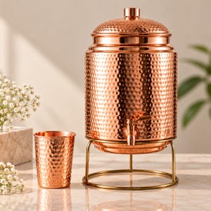 May include: A hammered copper water dispenser with a matching lid and a small spigot, resting on a gold-toned metal stand. Beside the dispenser sits a single hammered copper tumbler, both featuring a reflective, metallic finish and texture.