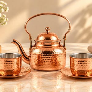 May include: A polished copper tea set consisting of a hammered teapot with a curved handle and spout, accompanied by two matching hammered cups and saucers. Each cup features a smooth silver-toned interior lining, contrasting with the exterior.