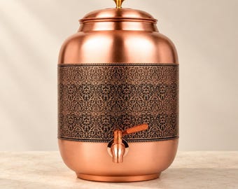 May include: A cylindrical copper water dispenser featuring a polished metallic finish and a central band decorated with a dark, intricate floral pattern. The vessel includes a matching copper lid with a small knob and a functional metal spigot.
