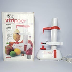 May include: A white and red Dazey Stripper electric fruit and vegetable peeler with a vertical post, a red top arm, and a base with a red power switch. The device is shown next to its original cardboard box featuring product text and illustrations.