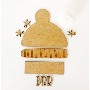 May include: A light brown wooden craft set featuring a beanie hat shape with a pom-pom cutout, a ribbed brim piece, a rectangular base, three snowflake-shaped accents, and the word BRR in cutout wooden letters for seasonal decoration projects.