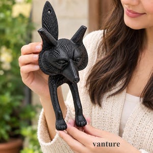 Könnte beinhalten: Ein schwarzer Wandhaken aus Gusseisen in Form eines Fuchses. Die Fuchsfigur weist eine detaillierte Fellstruktur, spitze Ohren und einen nach oben gerichteten Schwanz auf. Das Design umfasst ein Montageloch oben am Schwanz und zwei Vorderbeine.