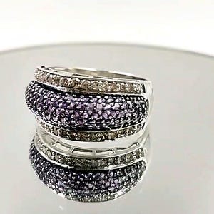 May include: A silver-toned ring featuring a wide, domed center band encrusted with numerous small purple stones, flanked by two thinner bands on the top and bottom edges that are set with rows of small, clear, sparkling white stones.