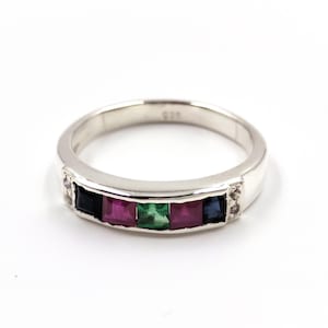 May include: A silver ring featuring a row of five square-cut gemstones in dark blue, magenta, green, magenta, and dark blue, flanked by two small clear stones on each side. The inner band is stamped with the number 925, indicating sterling silver.