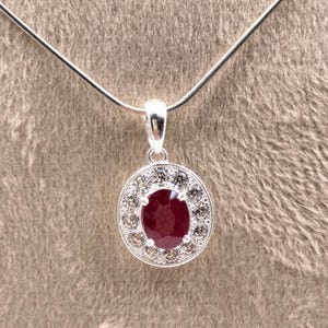 Puede incluir: Un collar con colgante de plata que presenta una piedra preciosa de color rojo intenso en forma ovalada, rodeada por un halo de pequeñas piedras claras de corte redondo. El colgante cuelga de una cadena de plata fina y lisa.