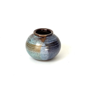 May include: A small, round ceramic vase with a glossy finish. The surface features a blend of mottled blue, green, and earthy brown tones with subtle horizontal ridges circling the body. The opening is circular with a dark brown rim.