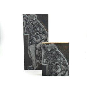 May include: Two rectangular black printing blocks featuring an engraved illustration of a musician playing a stringed instrument. The taller block shows the full figure, while the shorter block displays a cropped version of the same design.