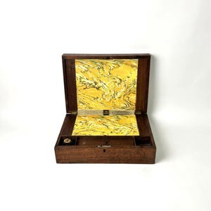 May include: A rectangular wooden box with a hinged lid, featuring a dark brown exterior finish. The interior surfaces of the lid and base are lined with a yellow and black marbled paper pattern. The base includes small compartments.