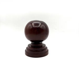 May include: A dark brown wooden finial featuring a smooth spherical top resting on a tiered, circular base. The object has a polished, reflective finish and a symmetrical design, standing upright as a standalone decorative component.