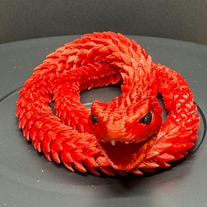 May include: A bright red articulated snake figurine with a segmented, scaly body texture. The snake is coiled in a circular position with its head facing forward, mouth open, and visible black eyes. The surface shows 3D printed layer lines.