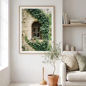 May include: A framed wall art print featuring a stone wall with an arched wooden window frame covered in lush green ivy. The image captures the textured masonry and the dense foliage surrounding the window, presented in a light wood frame.